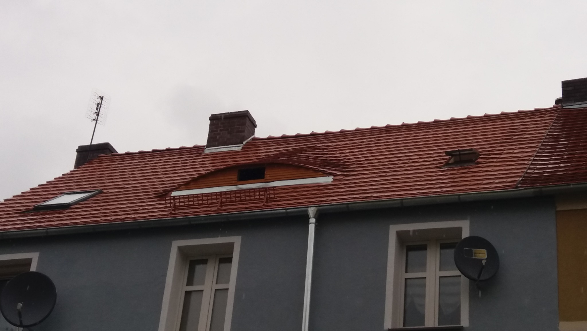 Czerwona dachówka na budynku z oknami dachowymi, kominami i anteną satelitarną, widok z dołu w pochmurny dzień.