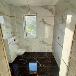 ProRemont - Wykończona łazienka z marmurowymi płytkami na ścianach i czarną podłogą. Widoczne białe sanitariaty i okno z widokiem na zieleń. Elementy wykończeniowe w kolorze złotym.