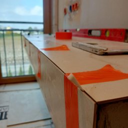 ProRemont - Wykończona płytkami zabudowa, zabezpieczona pomarańczową taśmą, z poziomicą w tle. Widok na okno i fragment instalacji elektrycznej.