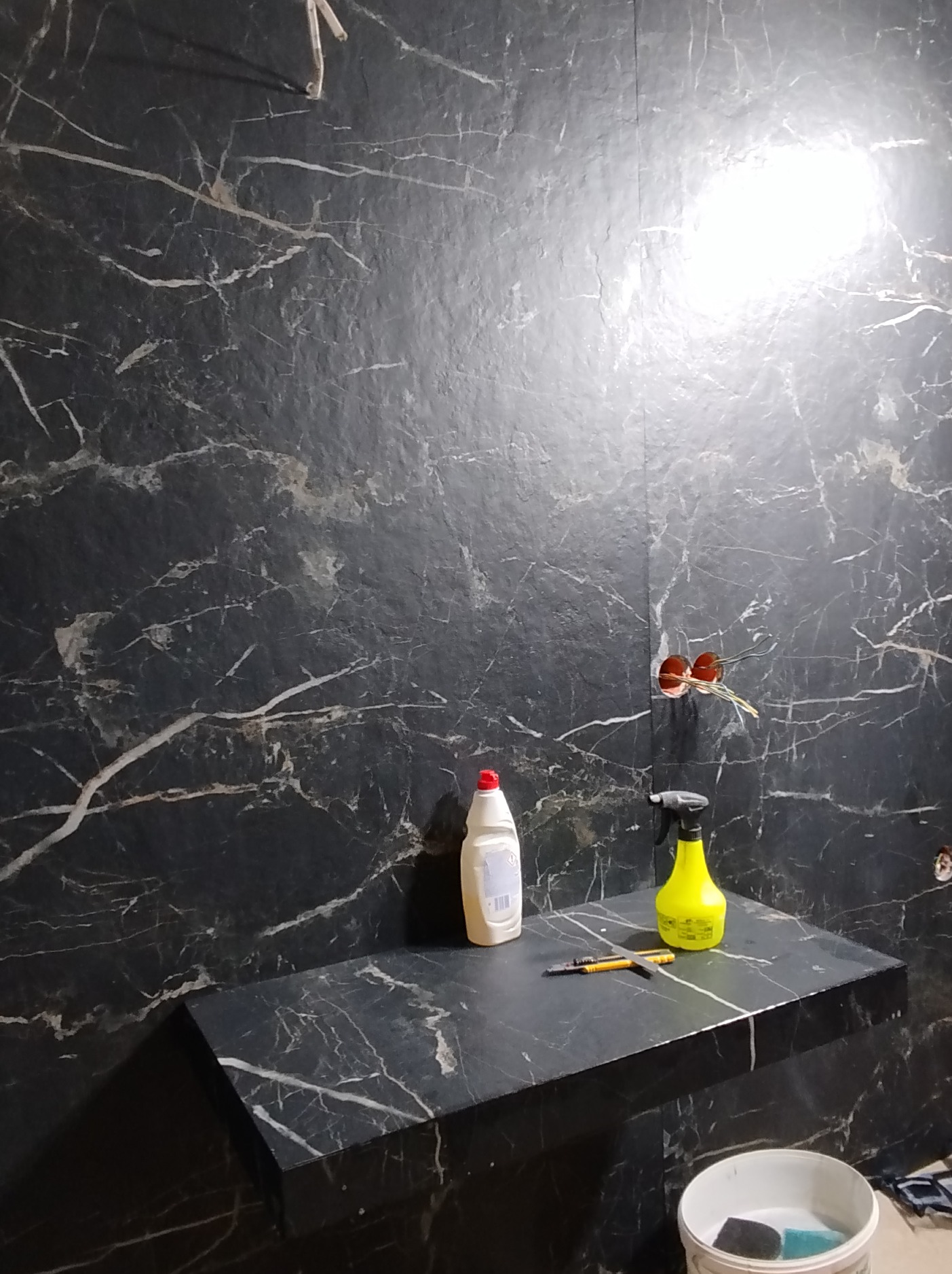 Wykończona ściana i blat z czarnego marmuru z białymi żyłami w łazience. Widoczne narzędzia i chemia budowlana. Gniazdka elektryczne bez osprzętu.