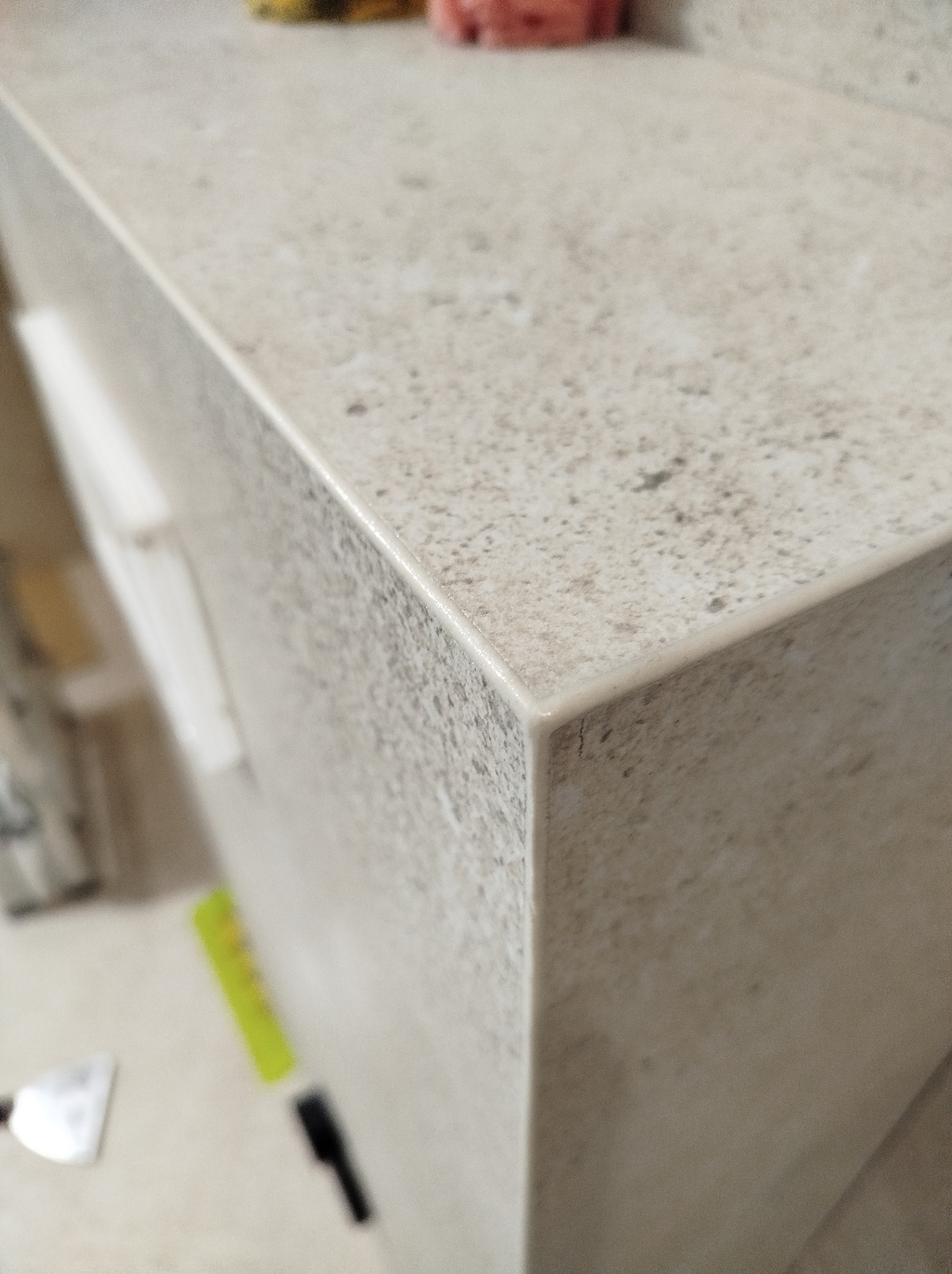 Wykończony blat łazienkowy z płytek w odcieniach beżu i szarości, z widocznym narożnikiem i fugą. W tle fragment szafki i narzędzia glazurnicze.