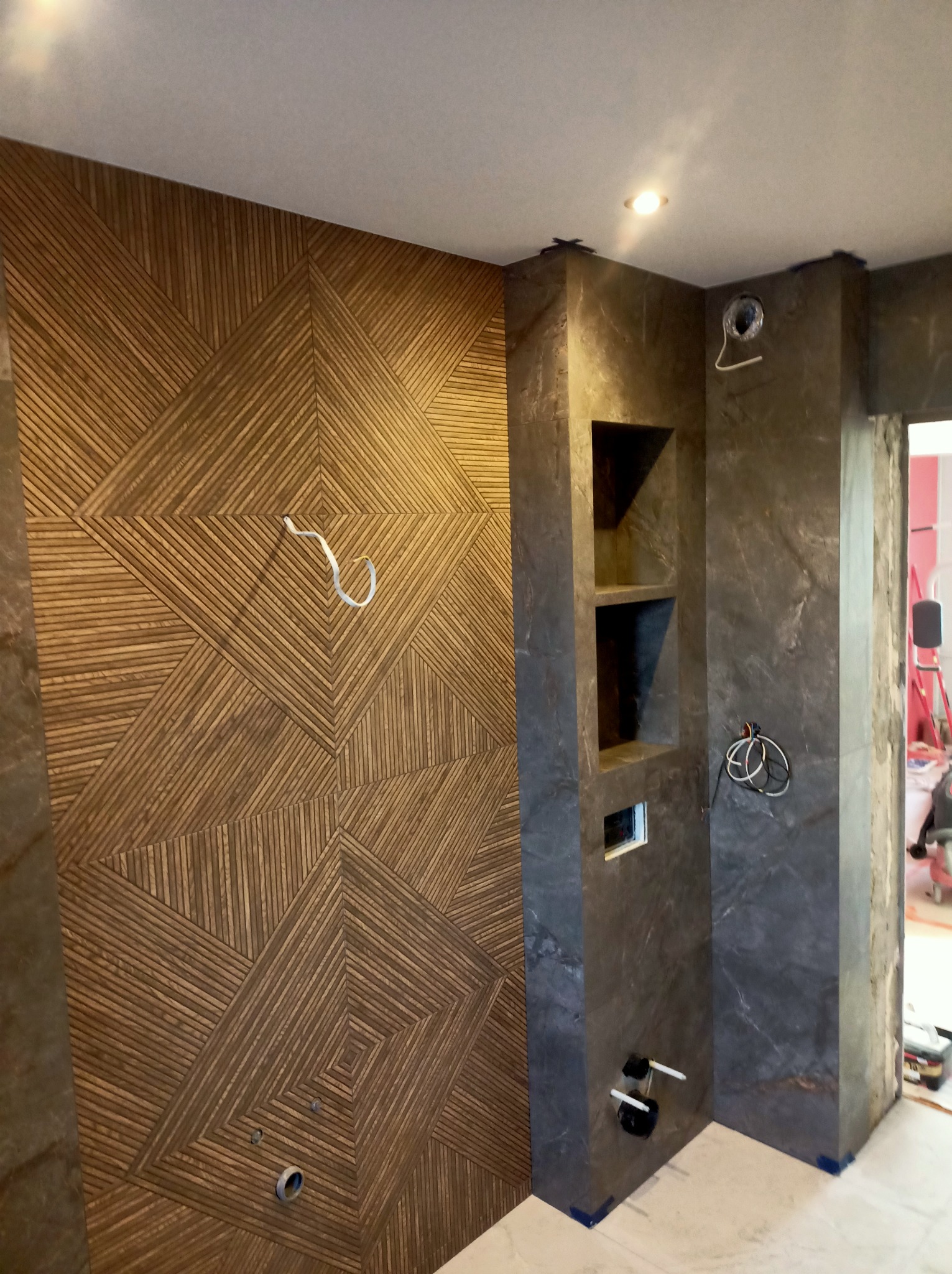 Wykończona ściana z drewnianą mozaiką w geometryczny wzór, obok ściana z szarego kamienia z wnękami i instalacją sanitarną. Nowoczesny design łazienki.