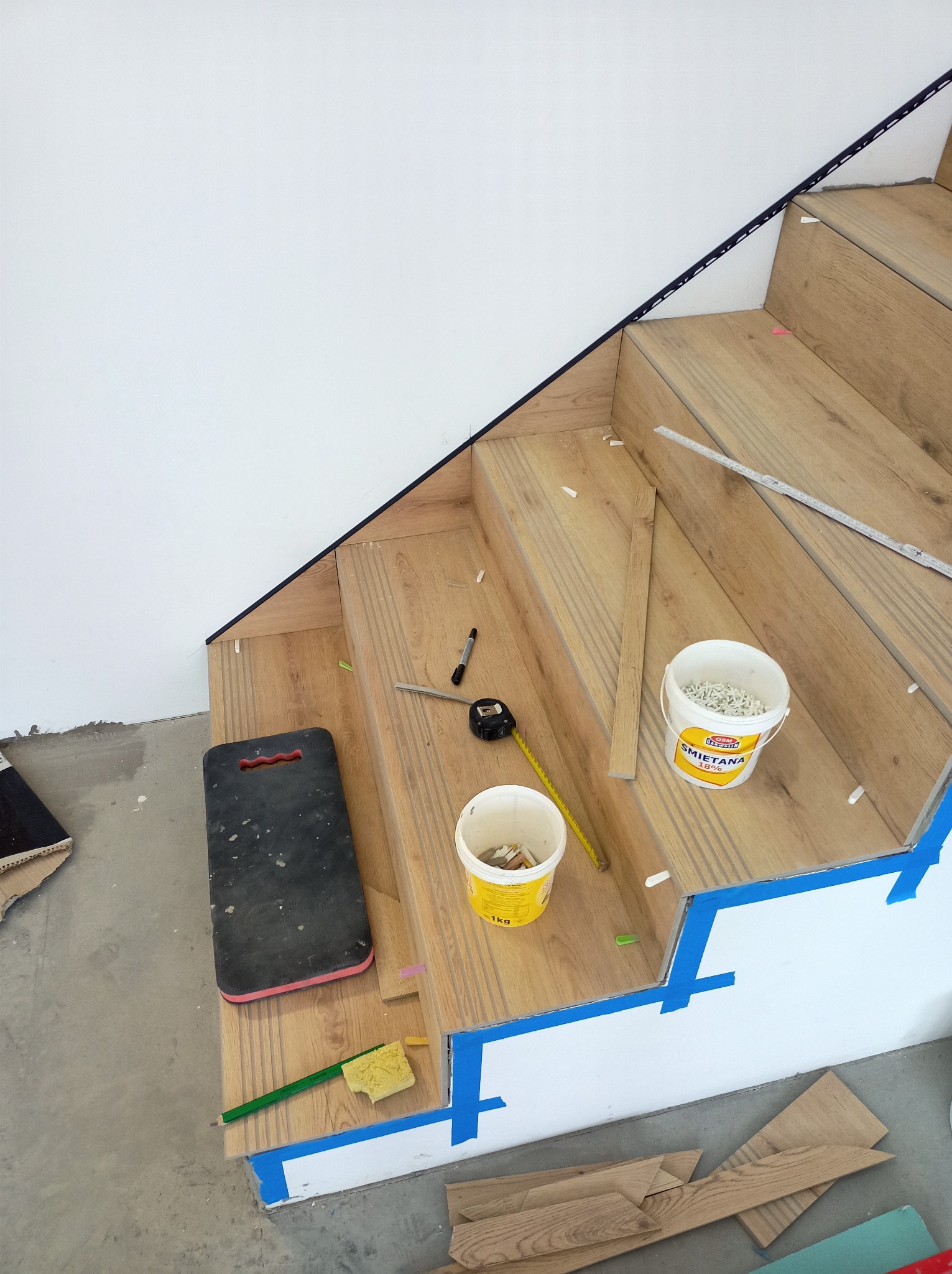 Wykończenie schodów drewnopodobnymi panelami. Widoczne narzędzia, klej i poziomica. Użycie taśmy malarskiej do zabezpieczenia krawędzi. Detal montażu paneli na schodach.