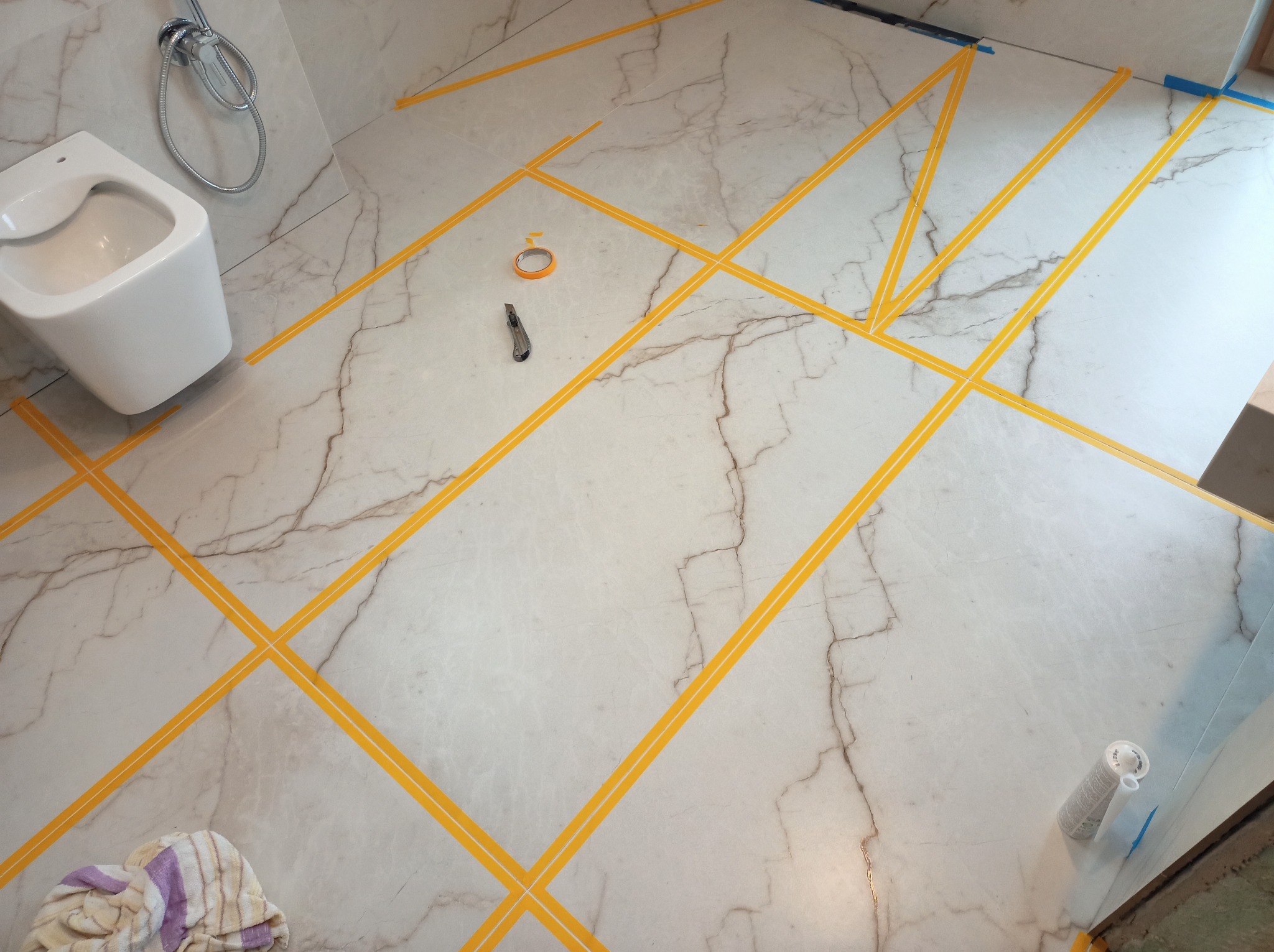 Biała marmurowa podłoga w łazience z wyznaczonymi żółtą taśmą liniami podziału płytek. Widoczny bidet, nóż i akcesoria glazurnicze.