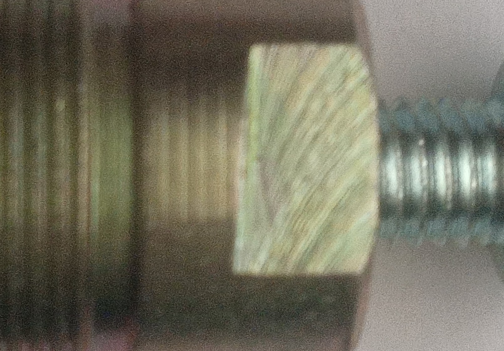 Szczegółowe zbliżenie gwintowanego metalowego pręta z kwadratową nakrętką, widoczne ślady obróbki skrawaniem na płaszczyznach nakrętki.