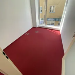 Pusty pokój biurowy z intensywnie czerwonym dywanem, widok na budynek z salonem SPA przez okno, białe ściany i drzwi, metalowe gniazda w podłodze.