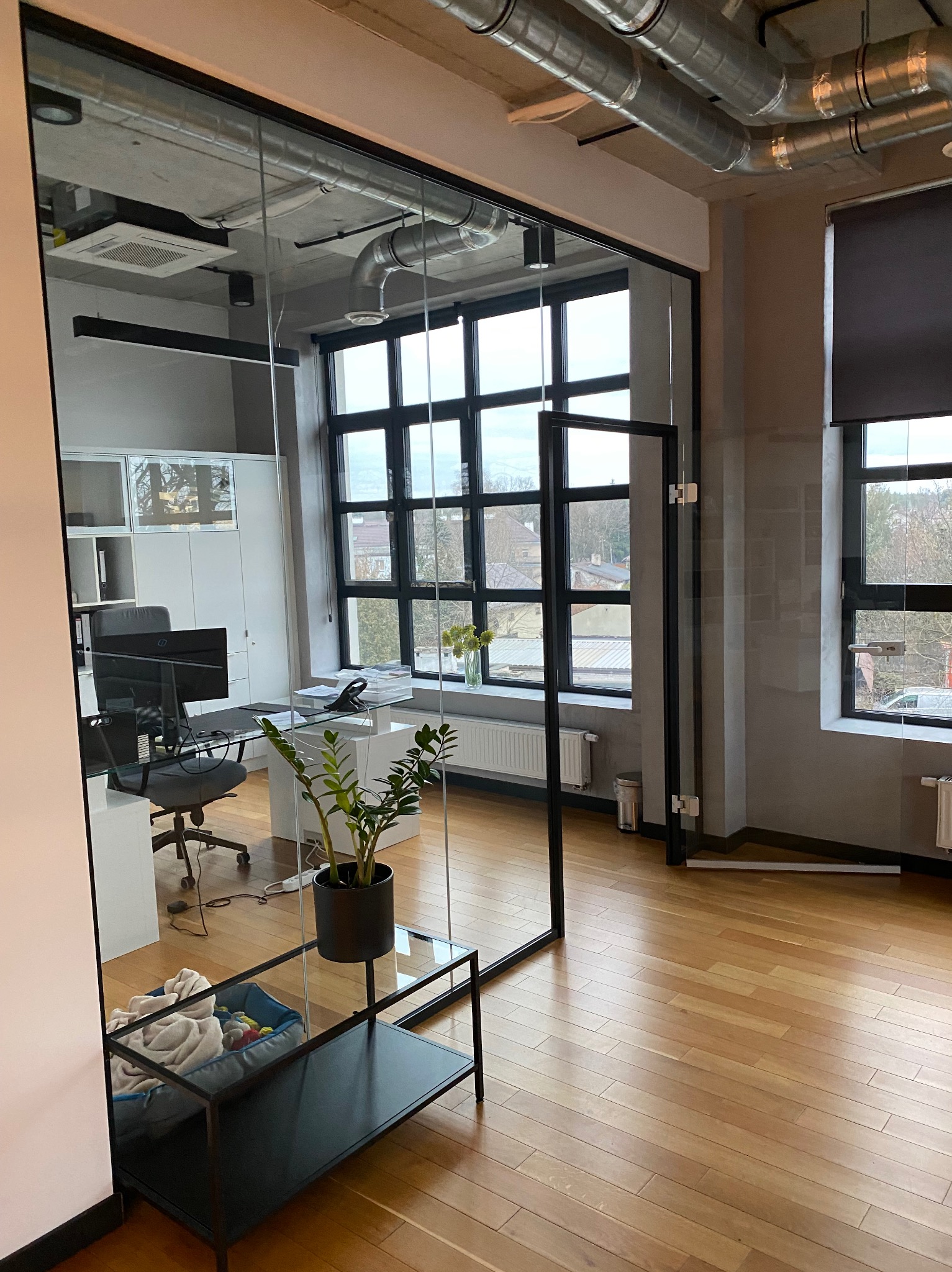 Nowoczesne biuro z przeszkloną ścianą działową, widoczne biurko, krzesło, roślina doniczkowa oraz elementy wentylacji pod sufitem.