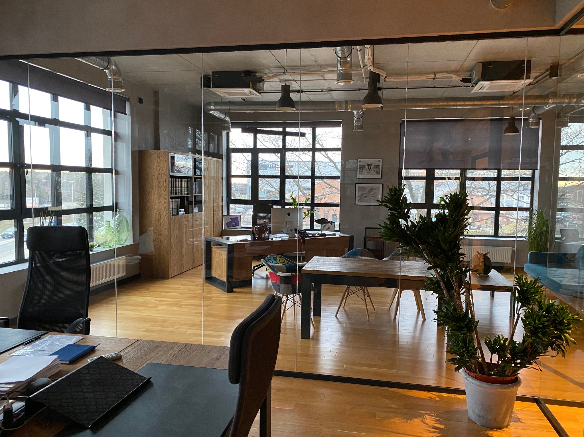 Nowoczesne biuro w stylu loft z drewnianą podłogą, dużymi oknami z widokiem na miasto i szklaną ścianą działową, oświetlone wiszącymi lampami i naturalnym światłem, z widocznymi elementami...