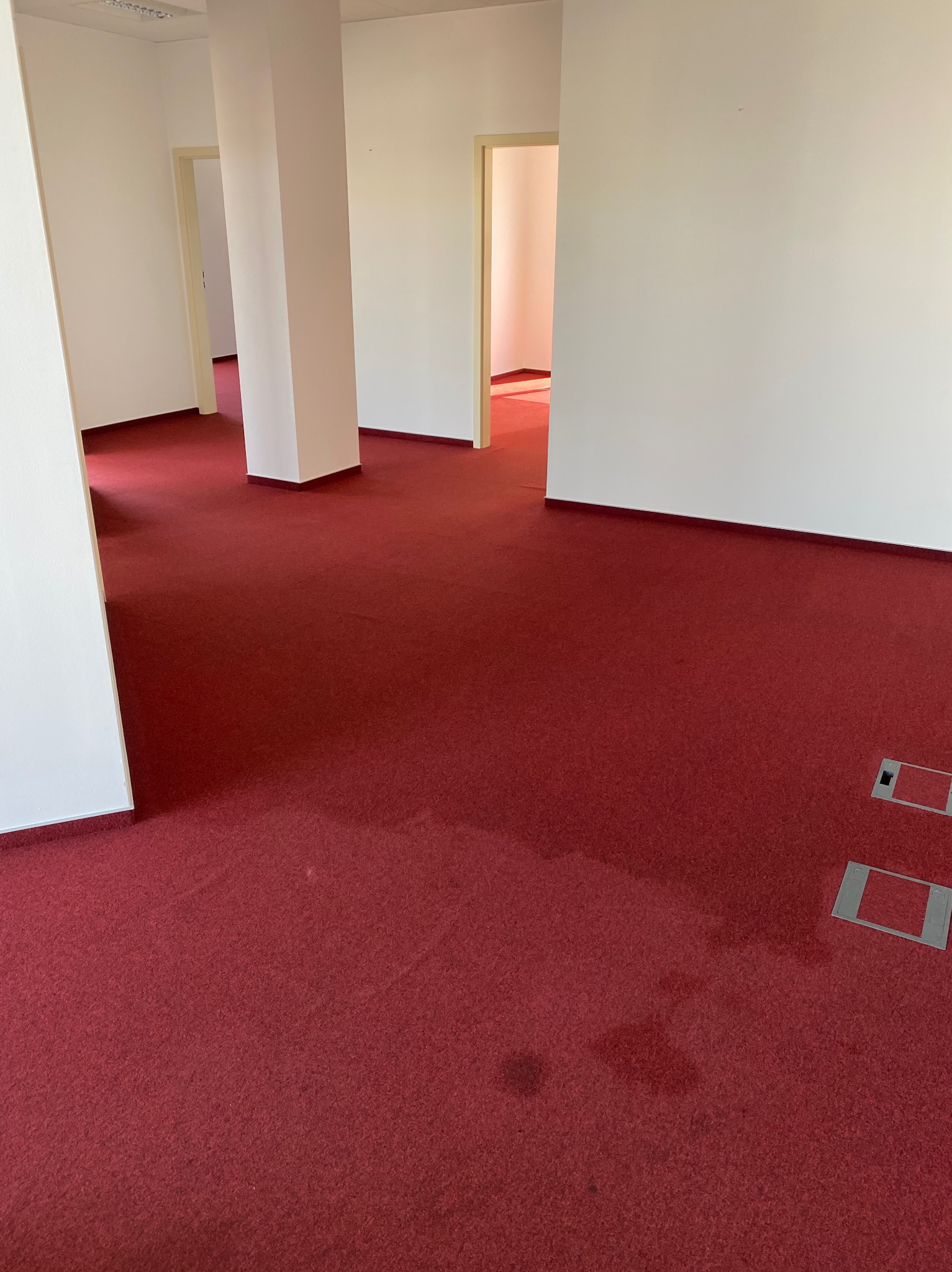 Czerwona wykładzina w pustym biurze z białymi ścianami i filarami, widoczne plamy na dywanie i gniazdka podłogowe.