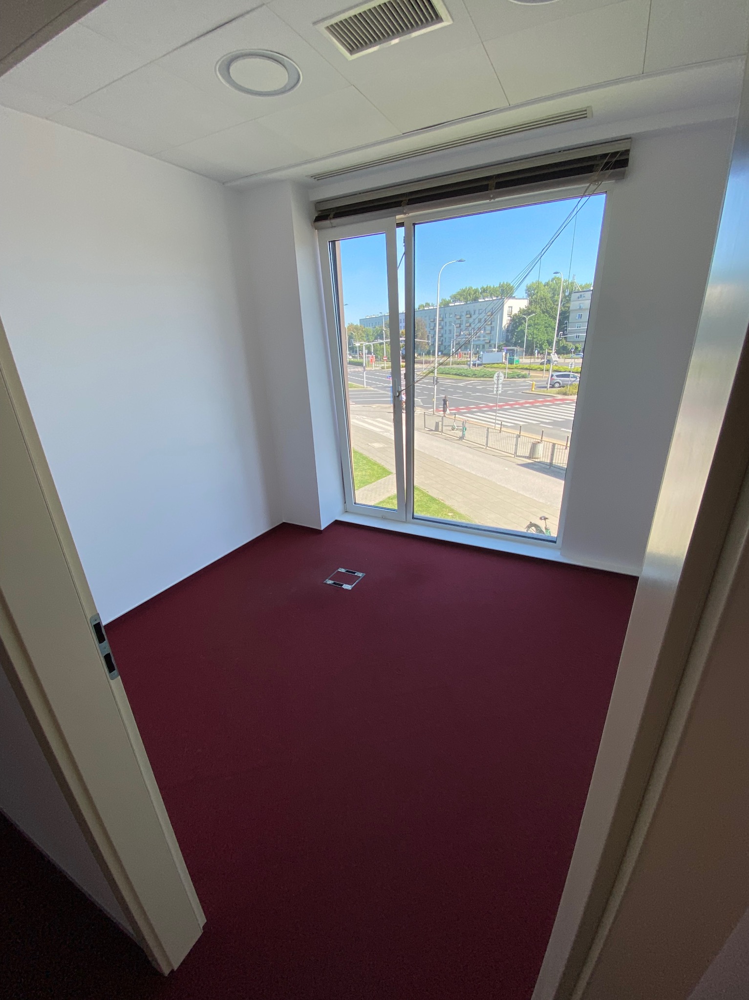 Puste biuro z burgundowym dywanem i widokiem na ulicę przez duże okno, widoczne kratki wentylacyjne na suficie.