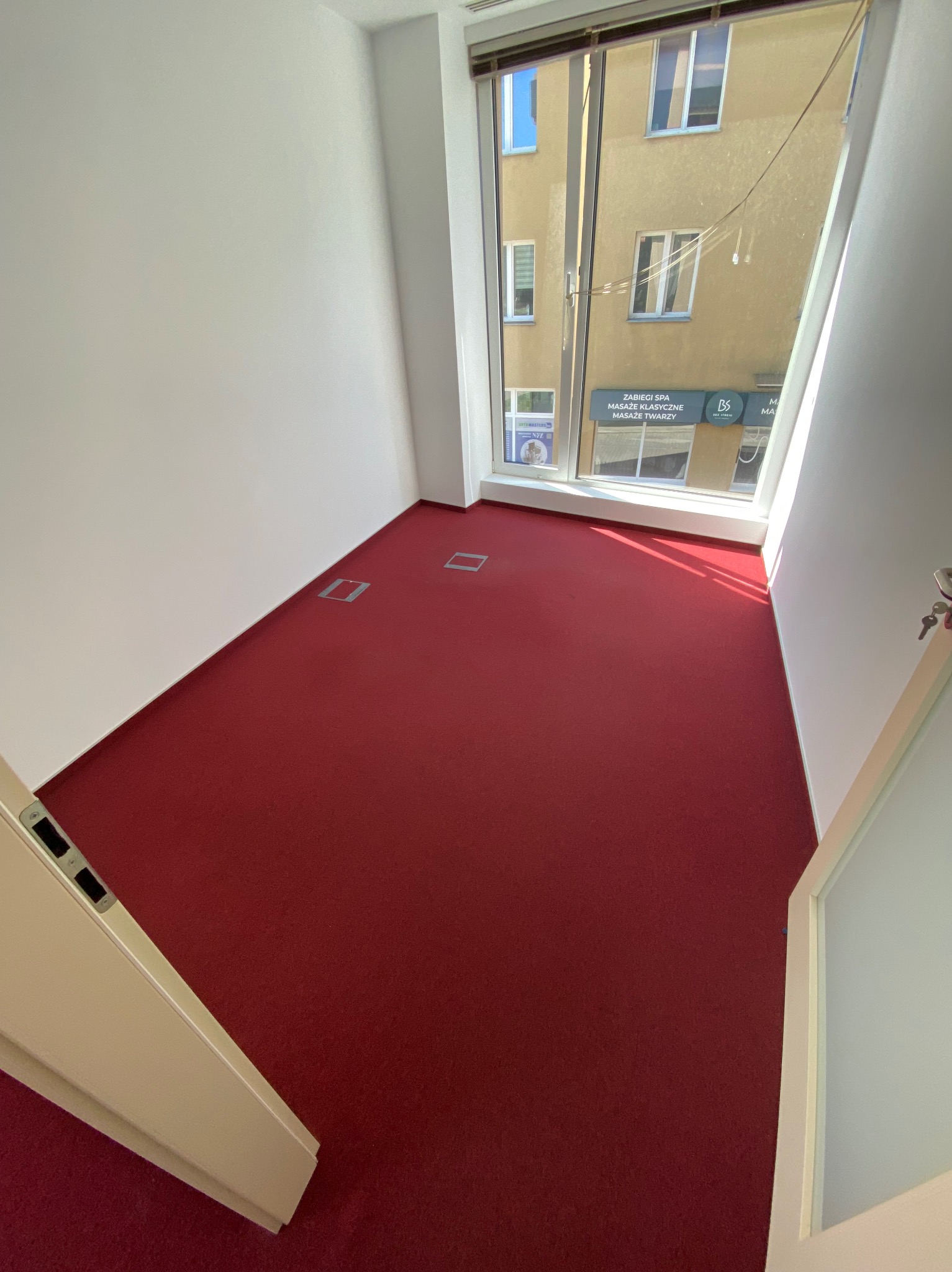 Pusty pokój biurowy z intensywnie czerwonym dywanem, widok na budynek z salonem SPA przez okno, białe ściany i drzwi, metalowe gniazda w podłodze.