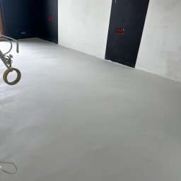 Paweł Kar - Świeżo wylana, gładka, szara posadzka w pomieszczeniu z czarno-białymi pasami na ścianach i wystającymi kablami. Na pierwszym planie widoczny fragment narzędzia z taśmą klejącą.