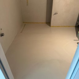 Paweł Kar - Świeżo wylana, jasnoszara posadzka w łazience, zabezpieczona taśmą malarską przy ścianach i odpływie liniowym, widok z góry.