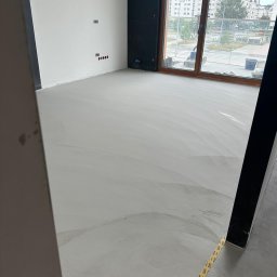 Paweł Kar - Świeżo wylana, jasnoszara podłoga w nowym mieszkaniu z widokiem na osiedle, widoczne ślady szpachlowania i taśma ochronna przy słupie.