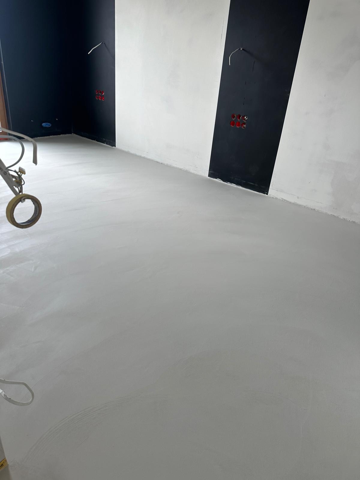 Świeżo wylana, gładka, szara posadzka w pomieszczeniu z czarno-białymi pasami na ścianach i wystającymi kablami. Na pierwszym planie widoczny fragment narzędzia z taśmą klejącą.