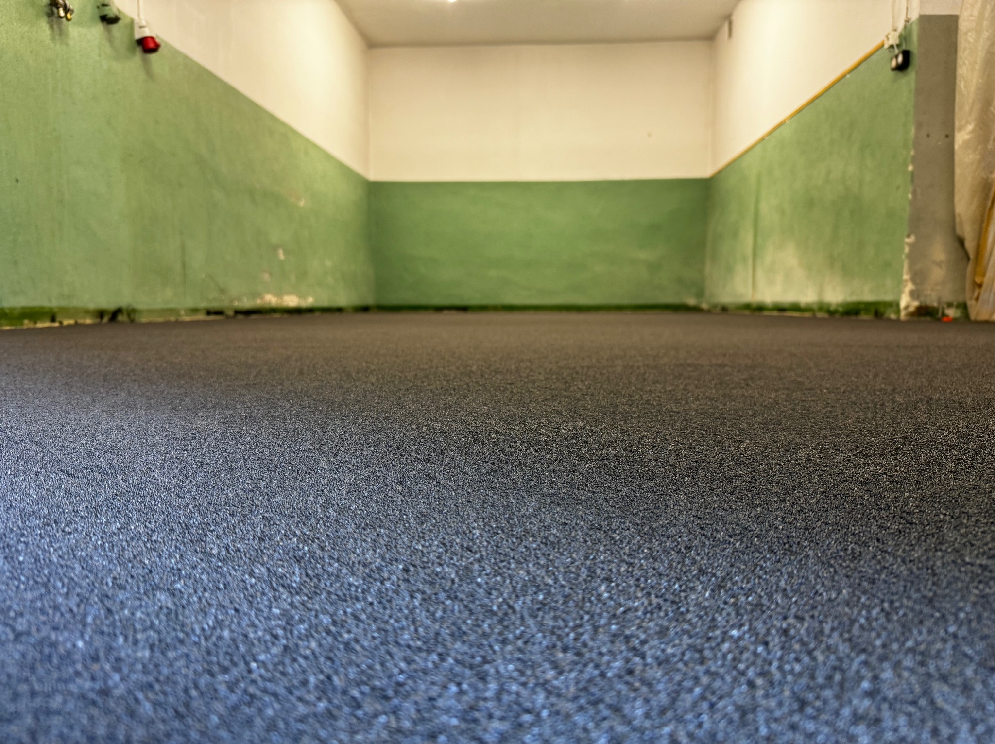 Nowa, szara posadzka żywiczna w hali przemysłowej, widok z poziomu podłogi, ściany pomalowane na zielono i biało, widoczne gniazdka elektryczne.