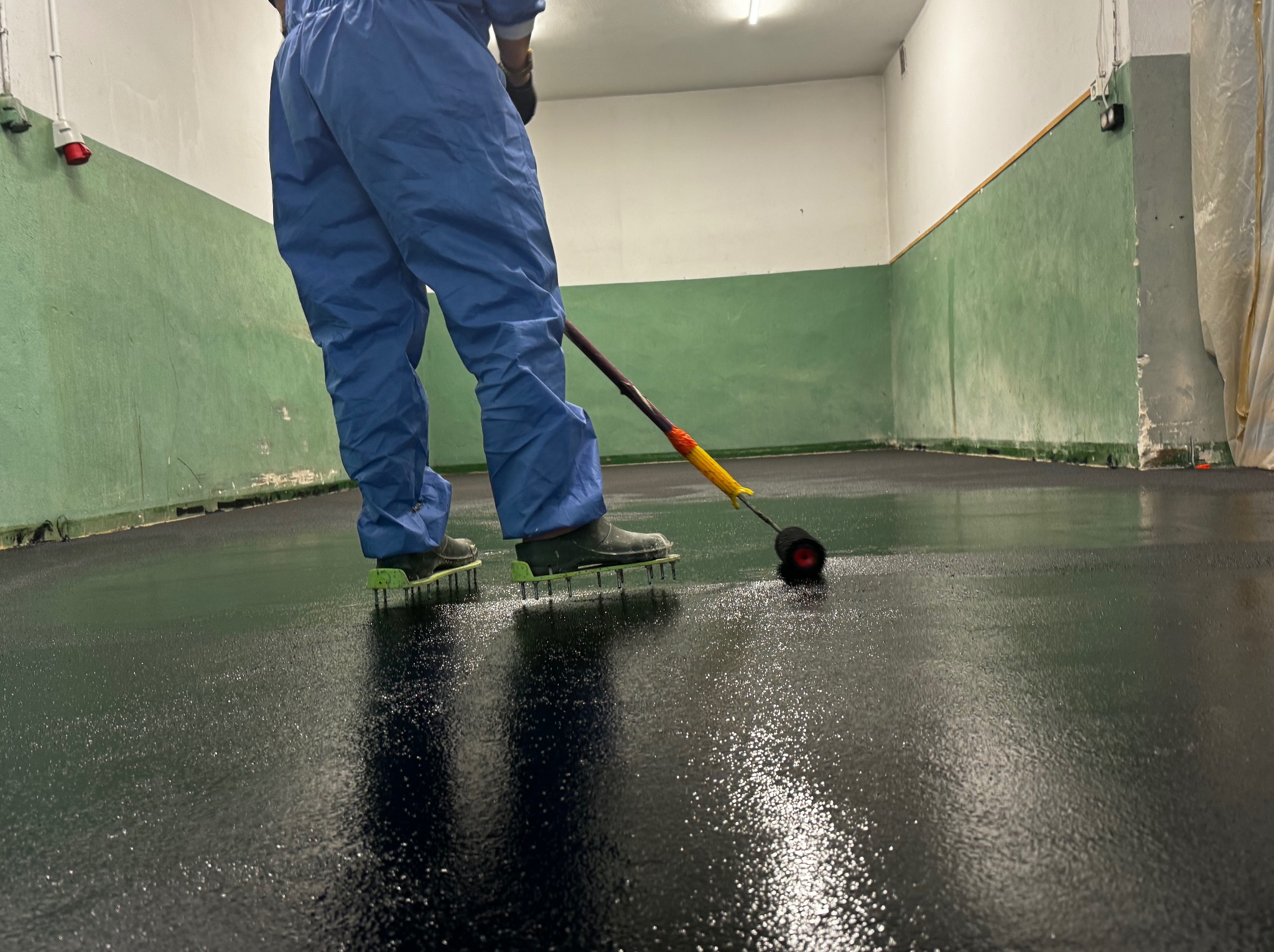 Pracownik w kombinezonie i kolcach na butach rozprowadza czarną, błyszczącą substancję wałkiem na długim kiju po podłodze w hali z zielono-białymi ścianami.