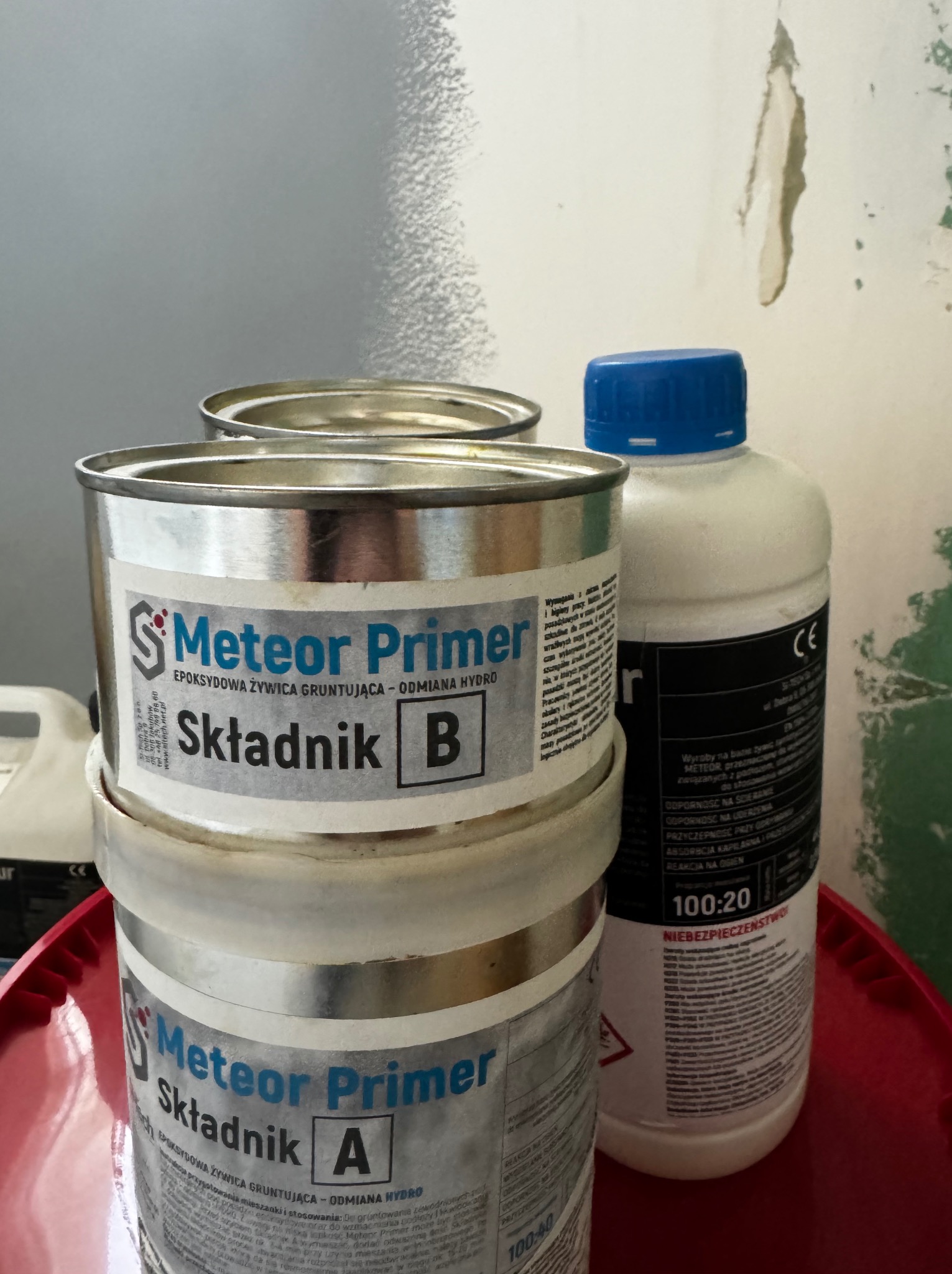 Dwa metalowe pojemniki z etykietami 'Meteor Primer, Składnik A' i 'Meteor Primer, Składnik B' oraz biała butelka z niebieską nakrętką na czerwonym podłożu, widoczne ślady uszkodzeń na ścianie w tle.