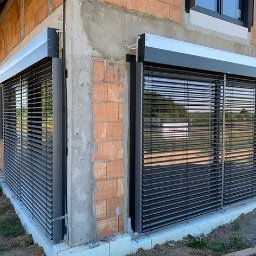 WINLUX Jakub Turowski - Grafitowe rolety wewnętrzne typu dzień noc zamontowane w oknach surowego budynku z czerwonej cegły, widoczne przewody elektryczne.