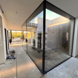 WINLUX Jakub Turowski - Wnętrze domu w trakcie wykańczania z narożnym oknem aluminiowym z widokiem na wewnętrzny ogródek skalny z drzewkami bonsai, widoczne narzędzia i kable na podłodze.