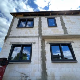 WINLUX Jakub Turowski - Surowy stan budynku z zamontowanymi oknami w czarnych ramach; widoczne białe bloczki, niedokończone spoiny oraz stos dachówek ceramicznych w lewym dolnym rogu.