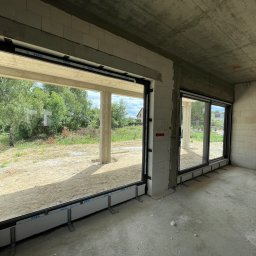WINLUX Jakub Turowski - Wnętrze surowego budynku z zamontowanymi dużymi oknami przesuwnymi PCV marki VEKA, widok na zewnątrz z zielenią i piaskiem, widoczne elementy montażowe.
