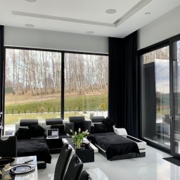 WINLUX Jakub Turowski - Nowoczesny salon w Szczecinie z dużymi oknami aluminiowymi z czarnymi ramami, w tle widok na drzewa, czarne zasłony i biała podłoga.