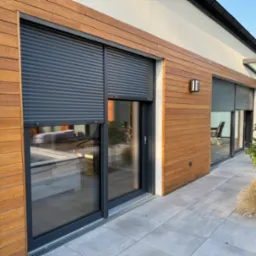 Zewnętrzny widok domu z drewnianą elewacją i oknami z zamontowanymi, częściowo opuszczonymi roletami zewnętrznymi w kolorze antracytowym.