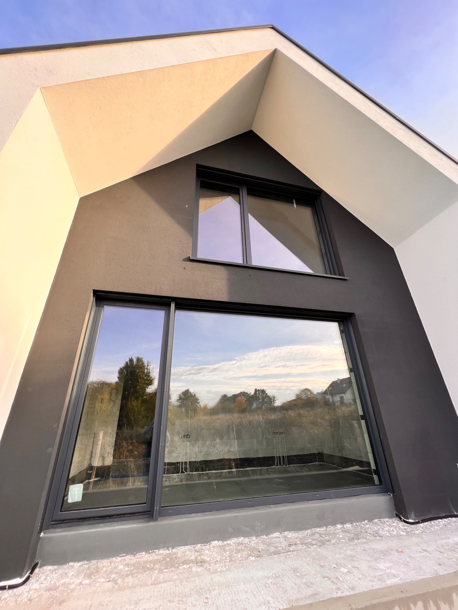 Nowoczesny dom z dużymi, grafitowymi oknami, widok z dołu na trójkątną fasadę, odbicie krajobrazu w szybach.