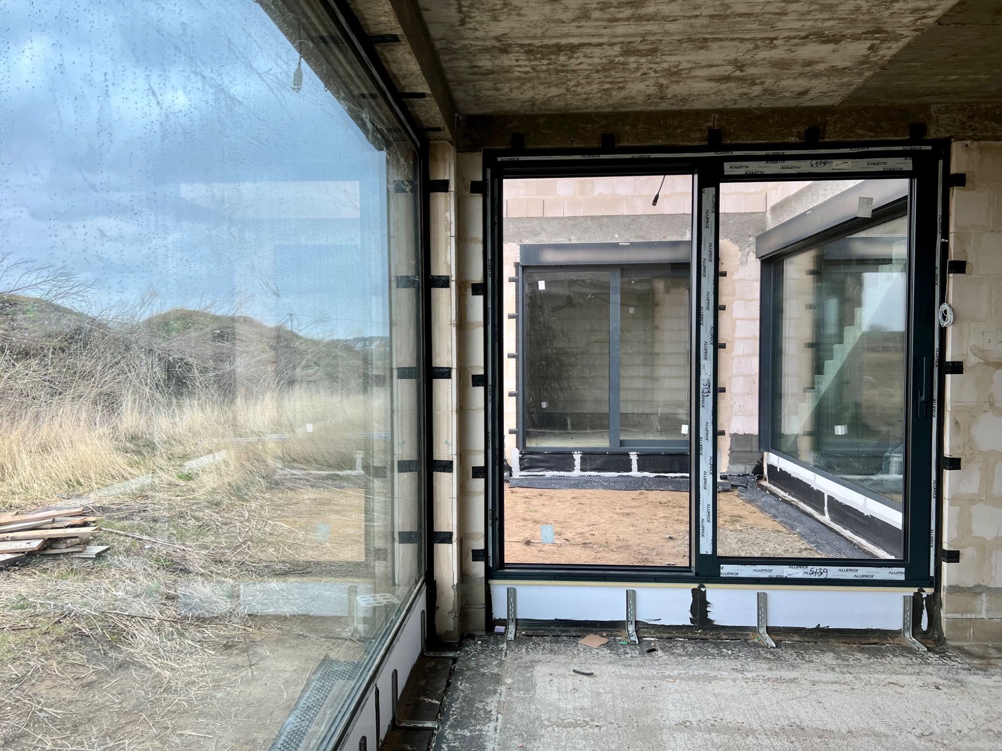 Surowy stan budynku z dużymi, panoramicznymi oknami z czarnymi ramami, widok na zewnątrz z polną roślinnością, widoczne elementy montażowe i surowy beton.