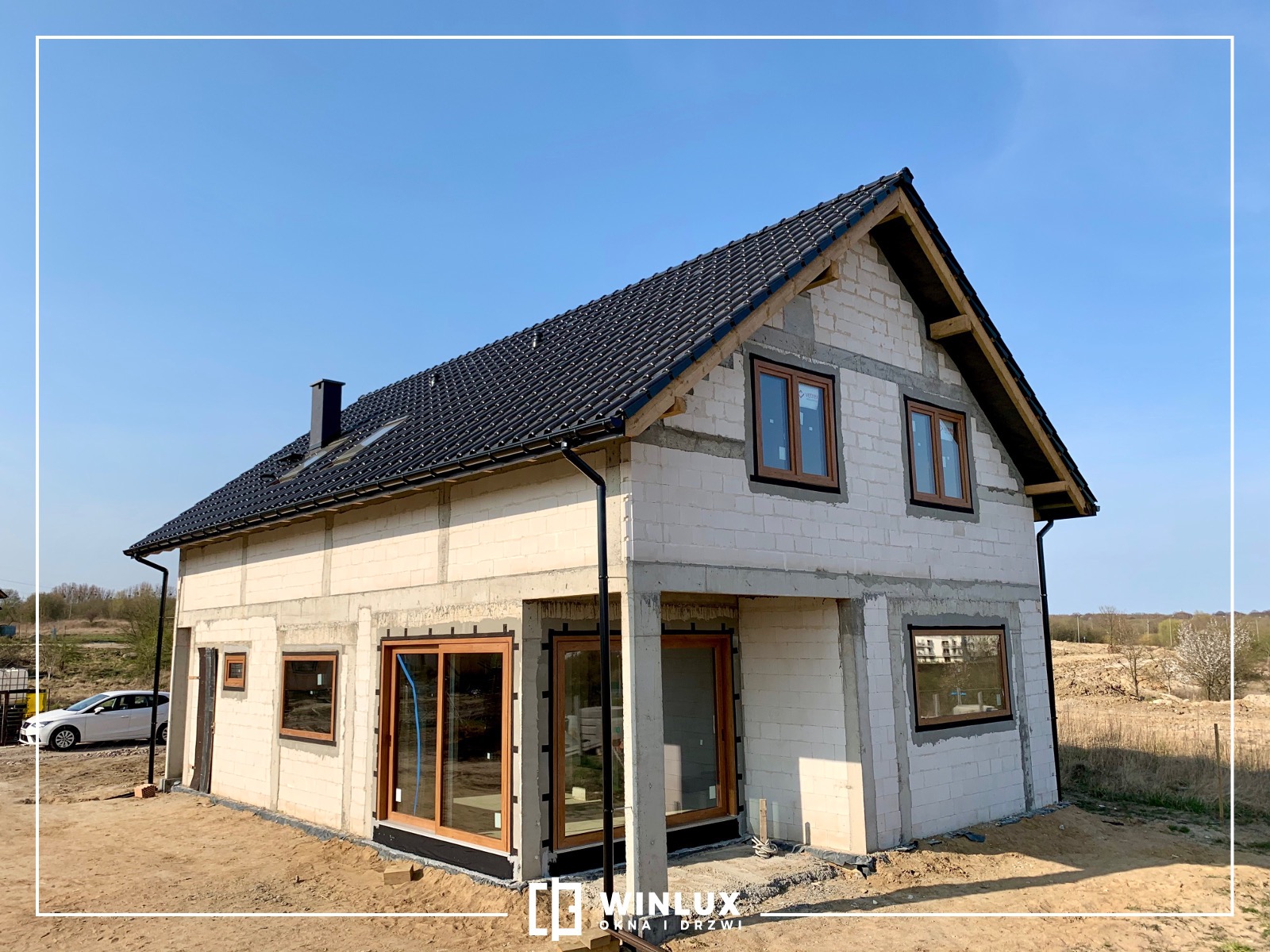 Budynek w trakcie budowy z wstawionymi oknami w drewnianych ramach i przygotowanym otworem na drzwi wejściowe, widoczne detale montażu okien.