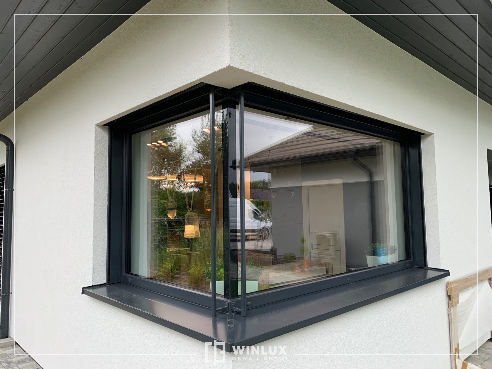 Narożne okno aluminiowe w kolorze antracytowym, z widocznymi elementami konstrukcyjnymi ramy, zamontowane w białej elewacji budynku, z odbiciem wnętrza domu.