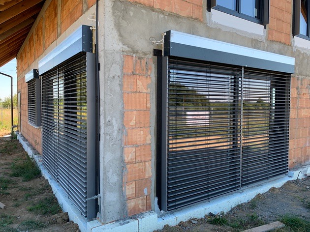 Grafitowe rolety wewnętrzne typu dzień noc zamontowane w oknach surowego budynku z czerwonej cegły, widoczne przewody elektryczne.