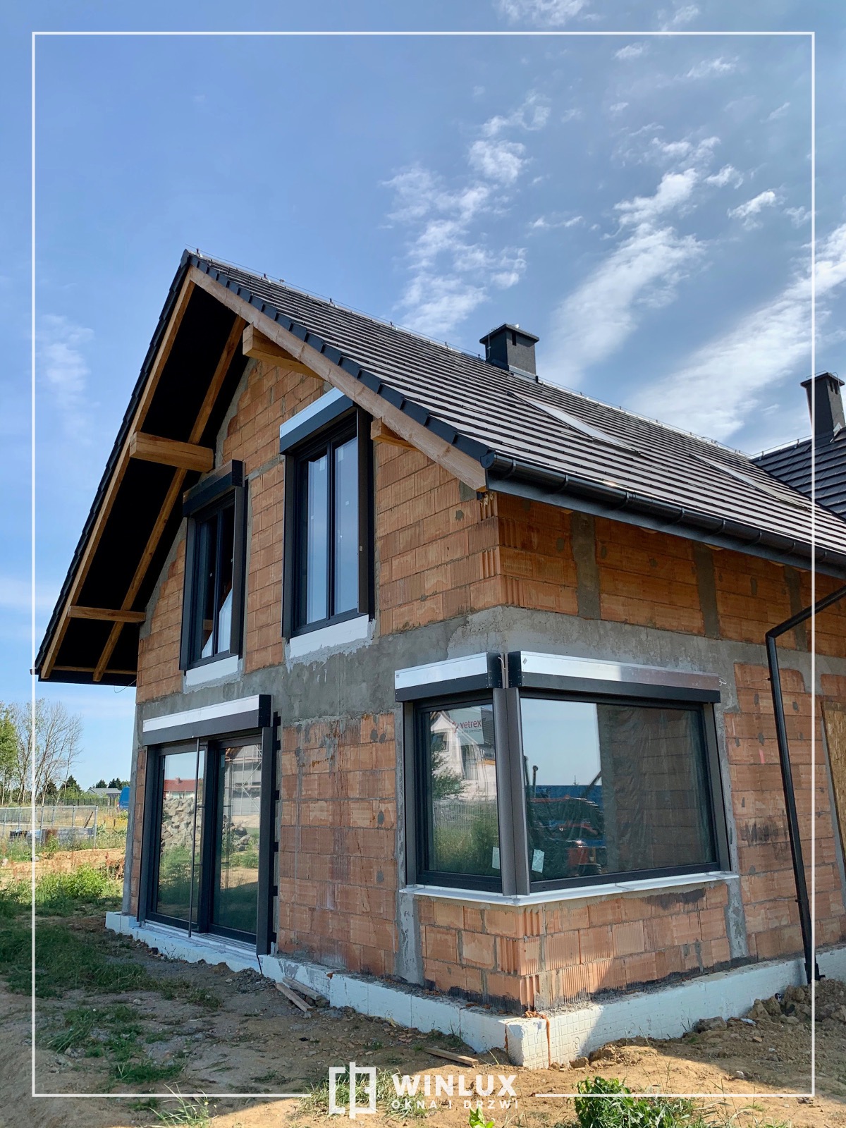 Surowy stan domu w trakcie budowy z zamontowanymi oknami z roletami zewnętrznymi, widoczne cegły, betonowe fundamenty i fragment logo firmy Winlux w rogu.