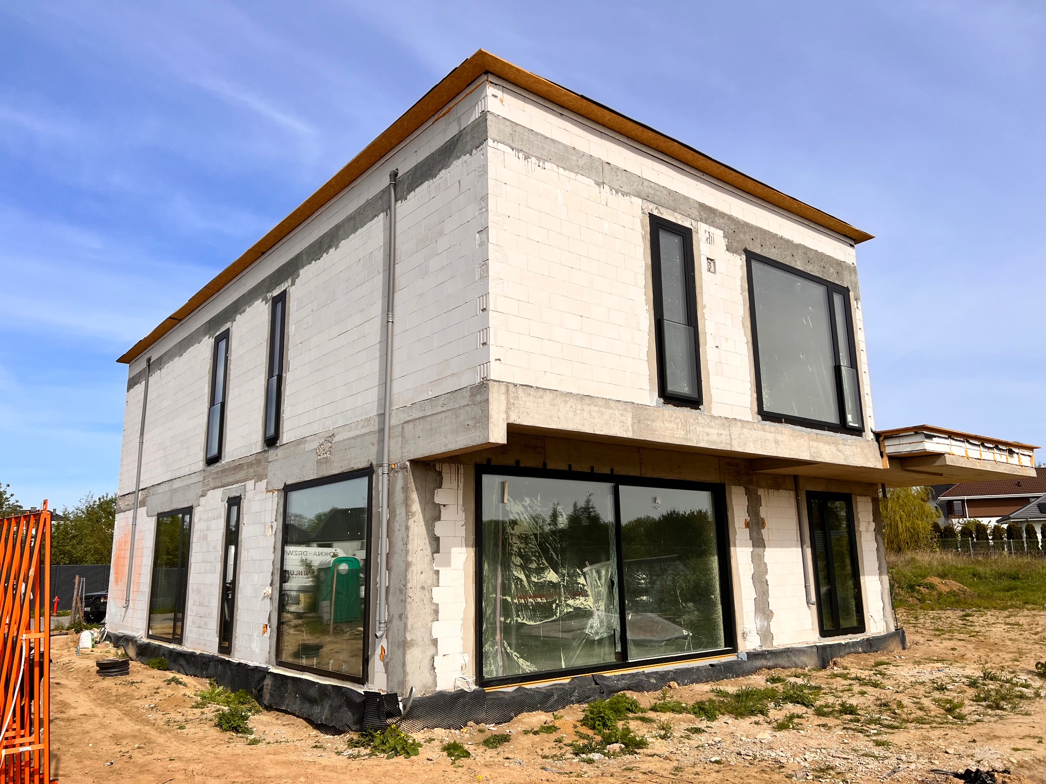 Surowy stan domu w trakcie budowy z zamontowanymi oknami z czarnymi ramami, zabezpieczonymi folią od wewnątrz, na tle błękitnego nieba.