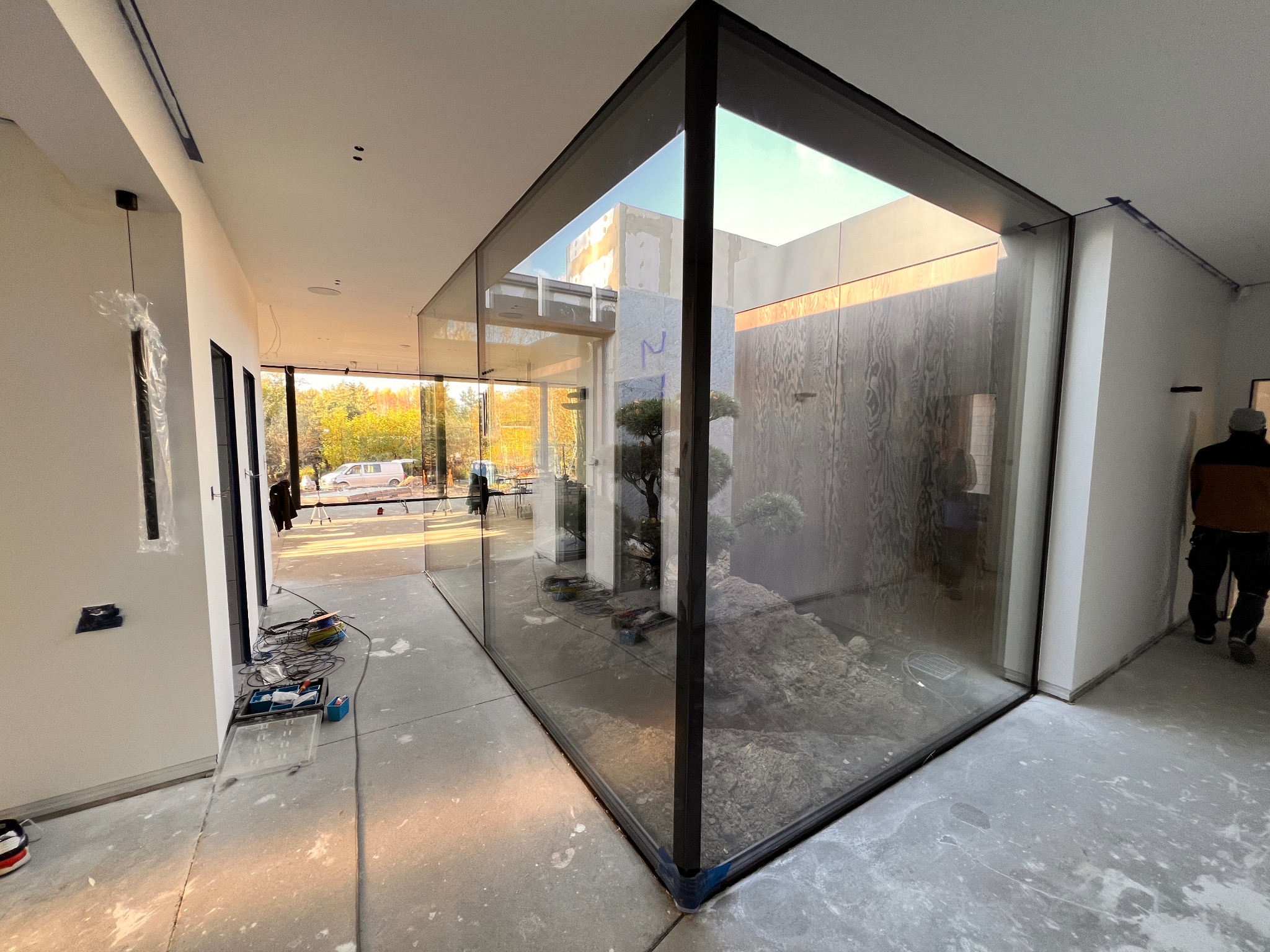 Wnętrze domu w trakcie wykańczania z narożnym oknem aluminiowym z widokiem na wewnętrzny ogródek skalny z drzewkami bonsai, widoczne narzędzia i kable na podłodze.