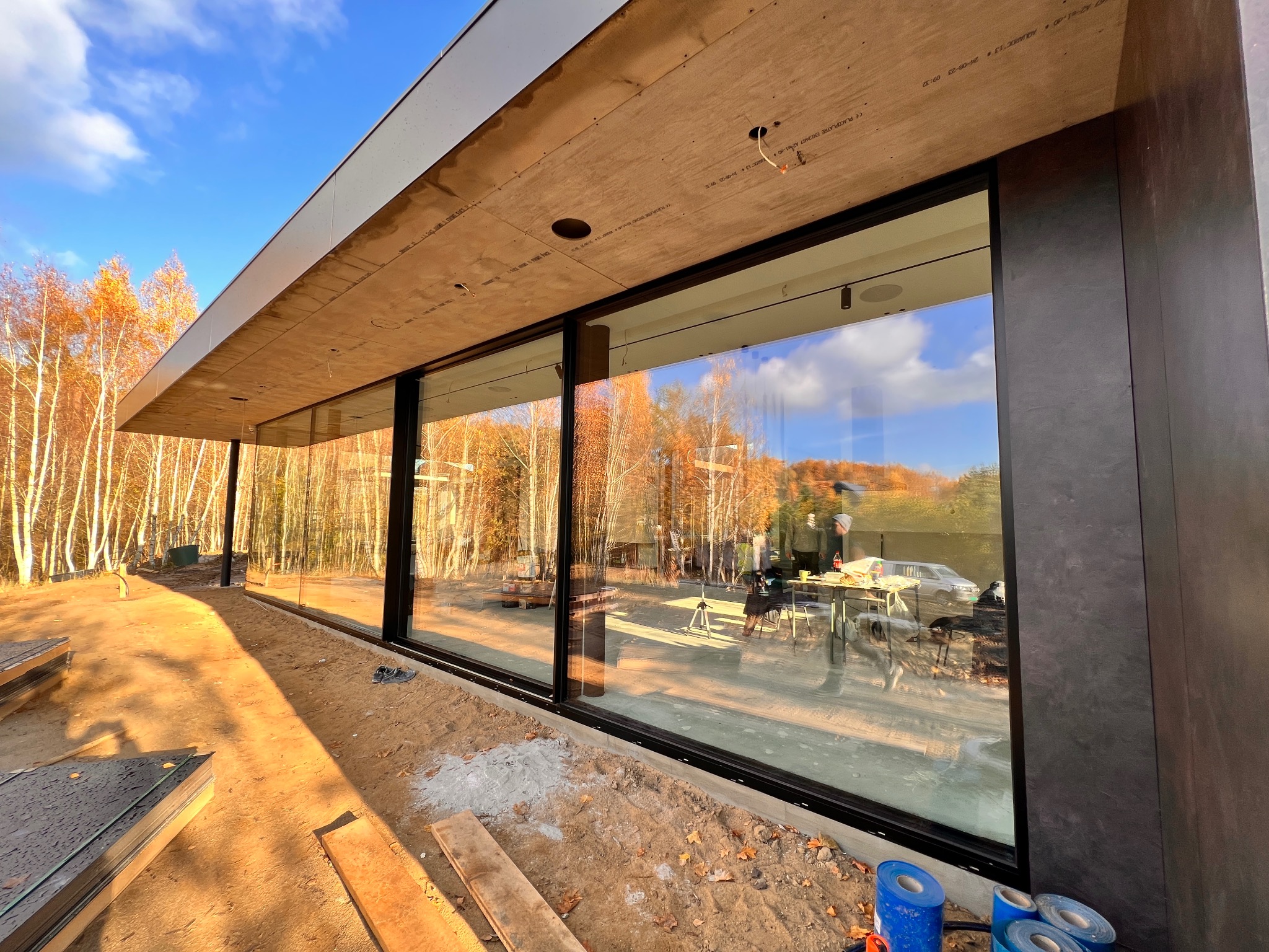 Nowoczesny dom w trakcie budowy z dużymi, panoramicznymi oknami z czarnymi ramami, odbijającymi jesienny krajobraz; widoczne materiały budowlane i narzędzia.