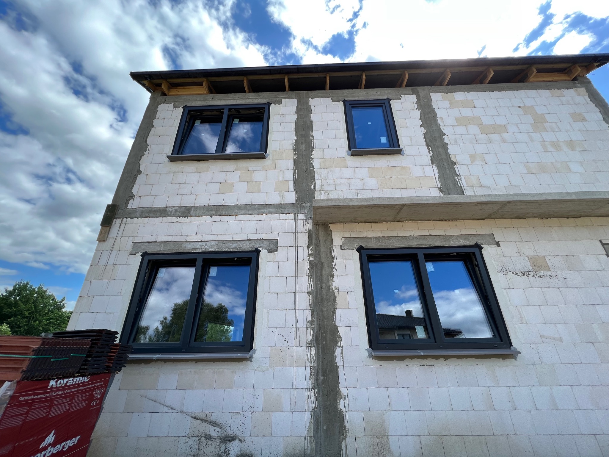 Surowy stan budynku z zamontowanymi oknami w czarnych ramach; widoczne białe bloczki, niedokończone spoiny oraz stos dachówek ceramicznych w lewym dolnym rogu.