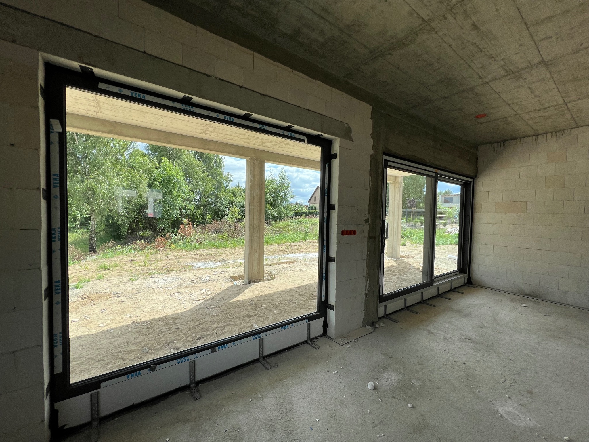 Wnętrze surowego budynku z zamontowanymi dużymi oknami przesuwnymi PCV marki VEKA, widok na zewnątrz z zielenią i piaskiem, widoczne elementy montażowe.