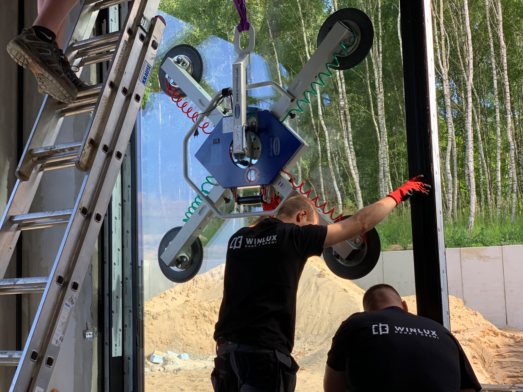 Montaż okna z użyciem przyssawki próżniowej; pracownicy w firmowych koszulkach Winlux, drabina Krause, widok na las brzozowy w tle.