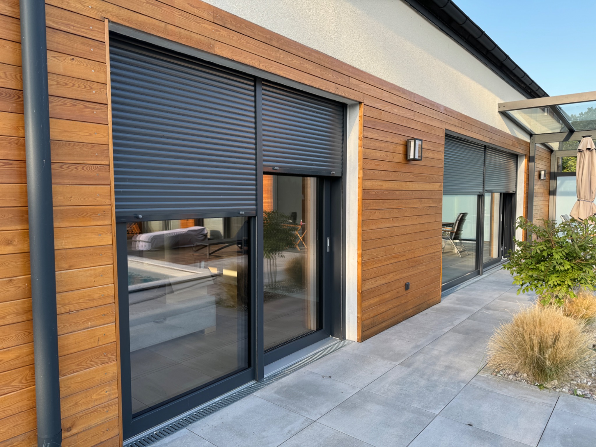 Zewnętrzny widok domu z drewnianą elewacją i oknami z zamontowanymi, częściowo opuszczonymi roletami zewnętrznymi w kolorze antracytowym.