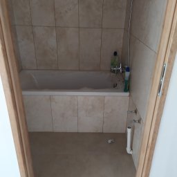 Usługi Remontowe Wykończenia Wnętrz - Wykończona łazienka z beżowymi płytkami, białą wanną, baterią i widocznymi rurami odpływowymi, sfotografowana zza drewnianej futryny.
