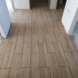 Usługi Remontowe Wykończenia Wnętrz - Ułożona podłoga z płytek imitujących drewno, widok z góry, surowe ściany i futryny w tle.