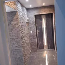 Korytarz z wykończonymi ścianami imitującymi kamień, szara podłoga z marmurkowym wzorem i nowoczesne drzwi wewnętrzne z pionowym, srebrnym paskiem, częściowo osłonięte folią ochronną.