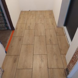 Usługi Remontowe Wykończenia Wnętrz - Ułożona podłoga z płytek imitujących drewno w wąskim przedpokoju, widoczne niedokończone obramowania drzwi i ślady po pracach budowlanych.