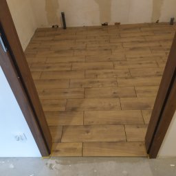 Usługi Remontowe Wykończenia Wnętrz - Układanie podłogi z płytek imitujących drewno w pomieszczeniu w trakcie prac wykończeniowych, widoczne niedoskonałości na ścianach i rurki instalacyjne, brązowa ościeżnica drzwiowa z żółtą taśmą...