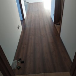 Usługi Remontowe Wykończenia Wnętrz - Korytarz z nową, ciemnobrązową podłogą z paneli, widoczne akcesoria montażowe przy drzwiach, świeżo pomalowane ściany na biało.