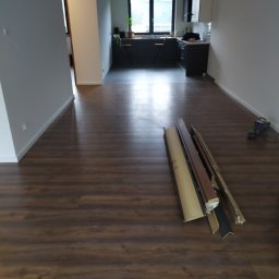 Usługi Remontowe Wykończenia Wnętrz - Przygotowanie do montażu paneli podłogowych w pustym pokoju z widoczną kuchnią w tle, narzędzia i listwy przypodłogowe leżą na podłodze.