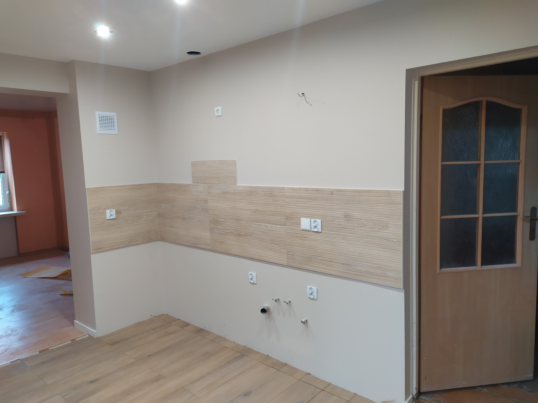 Fragment kuchni w trakcie prac wykończeniowych; częściowo położone płytki imitujące drewno na ścianie, widoczne gniazdka elektryczne, instalacja wodno-kanalizacyjna, drewnopodobna podłoga, otwór...