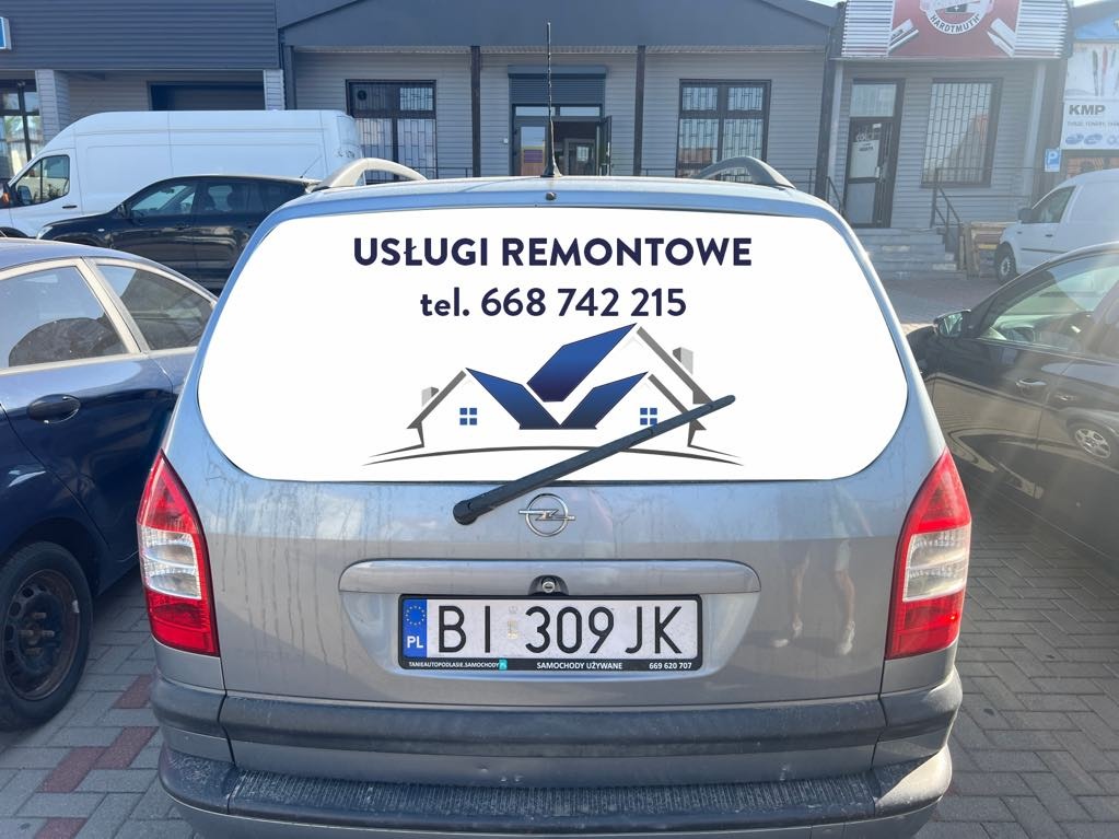 Tył srebrnego samochodu Opel Zafira z naklejką na tylnej szybie informującą o usługach remontowych, z numerem telefonu i graficznym logo firmy budowlanej.